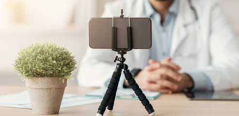 Telemedicine chat, virtual therapist consulting during social distancing. Friendly young arabian bearded male doctor in coat and glasses, look at smartphone webcam in office interior in clinic