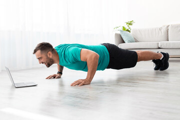 Cheerful millennial muscular european man doing push ups and watching online lesson on computer in living room interior. Workout and body care, new normal and sport during covid-19 lockdown, profile