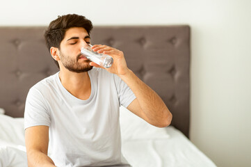 Starting new day with fresh aqua. Arab man taking glass of water sitting on bed in the morning,...
