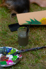 On the lush green grass, there rests a vibrant palette of colorful paint alongside a transparent glass filled with a liquid