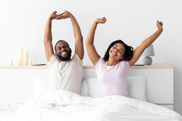 Happy millennial black husband and wife woke up, stretching bodies on white bed in minimalist...