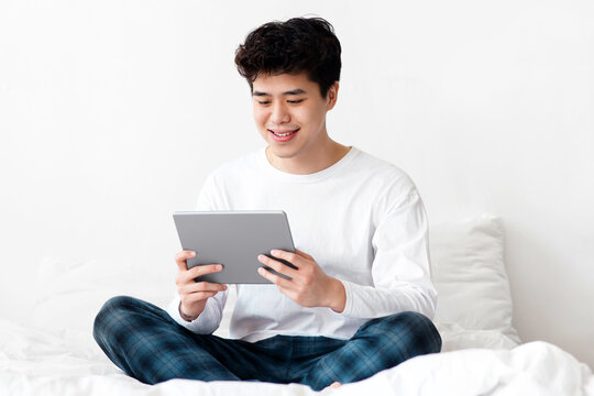 Modern device for homelander, education, online chat and work remotely. Cheerful asian young man in white t-shirt sits on bed in bedroom interior and looks at tablet after wake up, copy space