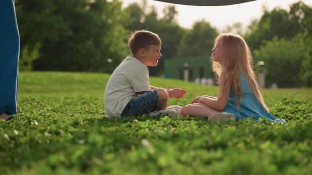 Playmates seated on grass while parents spreading blanket over them in sunny park, face painted girl and boy sharing joyful moment, green clover field background, family outdoor bonding