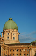 Obraz premium The majestic green dome of Buda Castle in Budapest stands proudly against a clear blue sky, with the subtle outline of the Fountain of King Matthias glimpsed below.