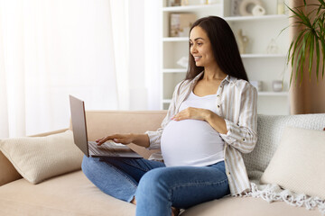 Smiling pregnant woman using laptop at home while holding belly. Casual maternity lifestyle...