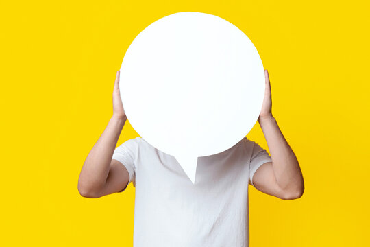 Unrecognizable man in jeans and white t-shirt hiding behind empty communication bubble over yellow studio background. Guy showing huge white blank clipart, copy space, communication, chatting concept