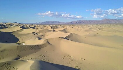 A sprawling desert landscape unfolds under a bright blue sky dotted with fluffy clouds. Gentle, sandy dunes ripple across the scene, fading into distant mountains on the horizon