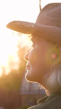 Sunset sunlight illuminates farmer woman profile wears straw hat. Portrait elderly female farmer on ranch. Warm rays of sun at sunset illuminate face. Person standing in rural landscapes