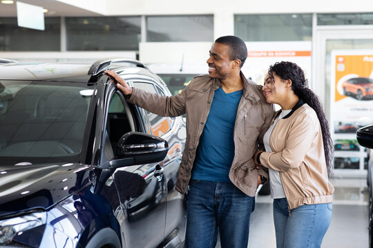 Young African American couple smiling while discussing new SUV inside dealership showroom, lifestyle and transport concept of auto sales, business and finance decision.