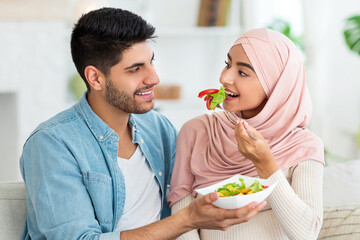 Caring arab man giving his young pregnant wife fresh vegetable salad, sitting on sofa in living room at home. Positive muslim couple eating healthy while waiting for baby