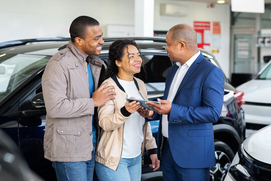 Young black couple with dealer looking at contract on tablet in car showroom, discussing purchase of new SUV, lifestyle and finance scene of modern auto shopping.