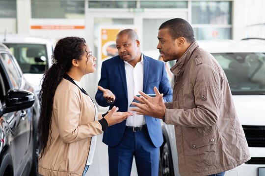 Young African American couple arguing in car showroom while dealer watches, conflict about purchase decision, lifestyle scene of disagreement and relationship stress