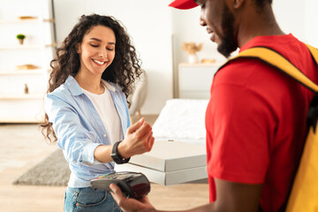 Fast Food Delivery, Easy Payment. African American male courier wearing red uniform holding Point of Sale terminal and pizza boxes, smiling female customer paying using wristwatch, standing at door