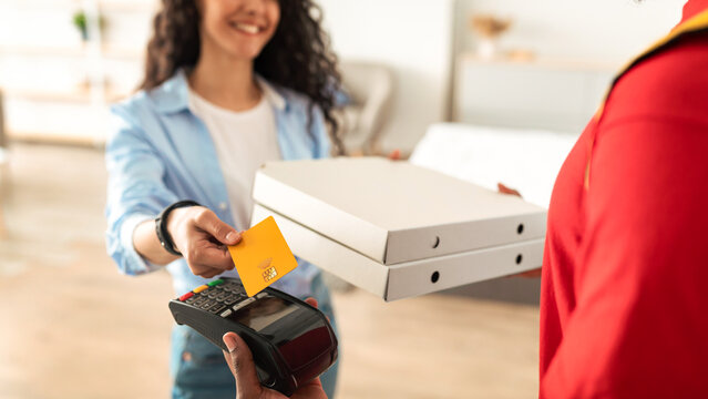 Delivery And Technology. Smiling woman using credit card for contactless payment, black male courier in red uniform holding POS machine in hand and pizza boxes to happy customer, selective focus