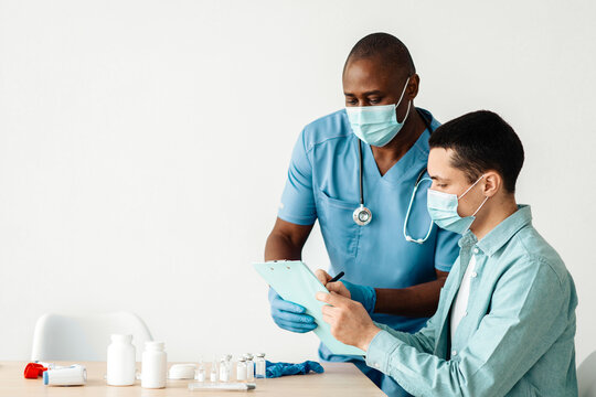 Preparation for immunization at covid-19 virus and vaccination procedure. African american millennial doctor in protective mask filling out form with patient at table with jars of drugs and syringes