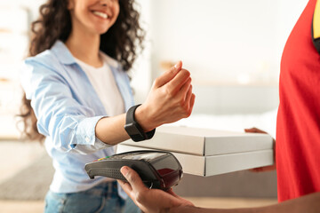 Contactless Financial Transaction Concept. Closeup of African American delivery guy holding payment terminal and carton pizza boxes, smiling woman paying using smartwatch, selective focus