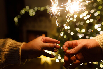Hands holding fireworks against christmas tree lights bokeh in evening cozy living room. Happy New...