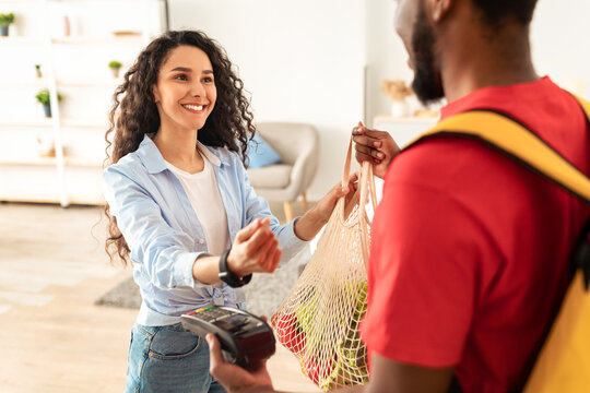 Grocery Shopping Delivery. Smiling Lady Paying With Smart Watch For Groceries Receiving Shopper With Food Products From Supermarket, Courier Guy Giving Mesh Bag To Client At Home. Selective Focus - Powered by Adobe