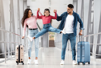 Fototapeta premium Cute Little Girl Having Fun With Parents While They Walking In Airport Together, Happy Arabic Family Enjoying Vacation Trip, Mom And Dad Holding Hands And Lifting Up Their Daughter, Copy Space