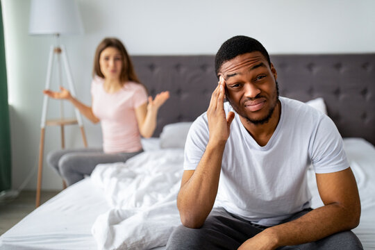 Displeased young guy sitting on bed, massaging temples in irritation, his angry wife shouting at him indoors. Millennial multiracial couple having marriage difficulties, facing relationship problems