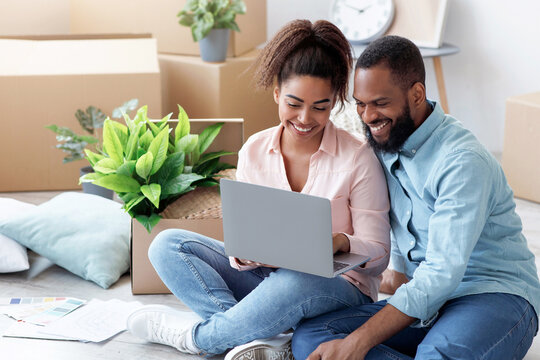 Smiling millennial african american wife showing to husband laptop with new room design, sitting on floor with boxes and plants. Couple planning interior of apartment, moving and buying furniture