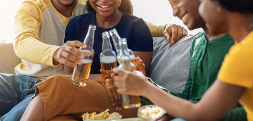 Group of black friends enjoying drinks party at home on weekend. Cheerful african american young men and women sitting in living room, cheering with beer bottles, home party concept, panorama