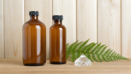 Two amber glass bottles placed on a wooden surface with a green fern leaf and decorative crystal against a natural wood background.
