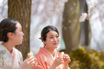 Obraz premium Two women in orange kimonos are eating sandwiches while sitting under a tree. Scene is relaxed and peaceful, as the women enjoy their meal in a serene outdoor setting