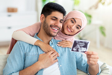 Loving muslim couple with ultrasound photo of their future baby resting at home on sofa. Positive...