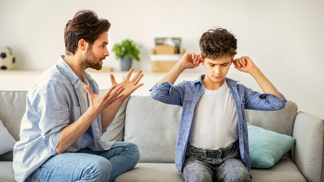 Parent violence. Angry dad scolding his innocent son, who covering ears not to listen angry father. Parent psychologically abusing boy, sitting together on sofa ta home
