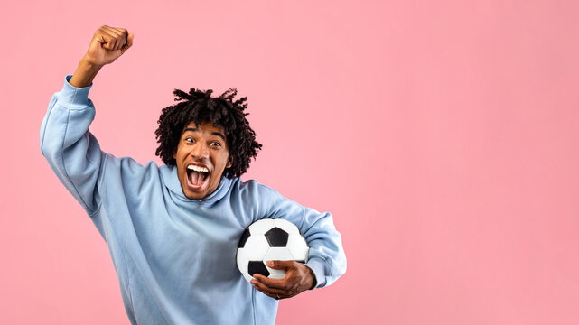 Happy black teenage guy with soccer ball rooting for his favorite football team on pink studio background. Excited African American sports fan celebrating victory, shouting GOAL