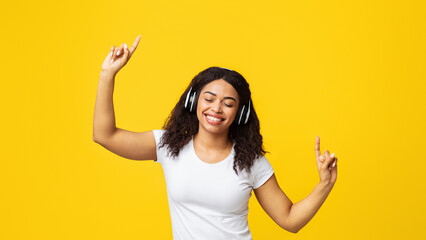 Move your body. Positive african american woman listening to music in wireless headphones with closed eyes, dancing and pointing up, enjoying new playlist over yellow background