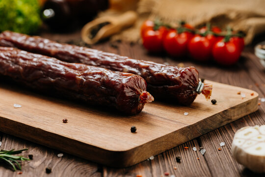 Dry cured sausage sticks on wooden board with herbs, tomatoes, and spices. Rustic food styling