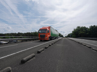 Orange truck with green trailer on highway with concrete barriers and overcast sky