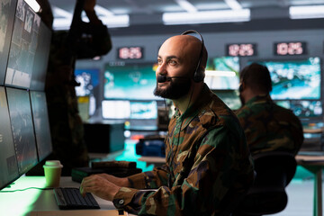 Army dispatcher looking at surveillance transmission on displays, relaying data to troops using...