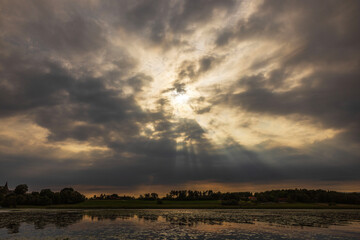 Dramatic sunset with sun rays breaking through dark clouds over calm lake surface. Sweden.