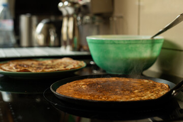 A large golden-brown blin fries on a skillet, with ready blini and batter, perfect for a family meal. Ideal for food visuals, home cooking, or cozy family lifestyle content with a warm appeal.