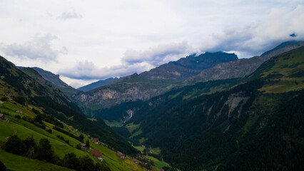 Obraz premium Swiss Alps with houses, winding road, clouds, and overcast sky, captured by drone. Schweizer Alpen mit Häusern, kurviger Straße, Wolken und bedecktem Himmel, aufgenommen mit Drohne.