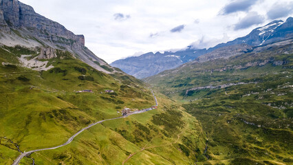 Naklejka premium Swiss Alps with houses, winding road, clouds, and overcast sky, captured by drone. Schweizer Alpen mit Häusern, kurviger Straße, Wolken und bedecktem Himmel, aufgenommen mit Drohne.