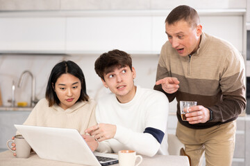 Family of adult man and young couple woman and guy sitting at laptop in kitchen..