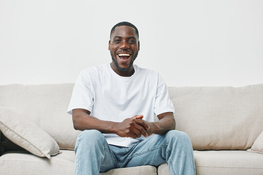 Happy African American man sitting on a couch in casual attire, expressing joy and positivity in a bright, minimalist living room setting