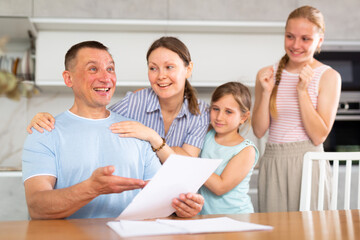 Happy adult man reading notification from employer informing of promotion or awarding bonus for achievements. Friendly family, wife and teenage daughters rejoicing together with father