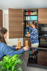Mother and daughter adding cookies packages to 72 hours survival kit emergency, ready for potential disasters, war or food shortage. Family cooperating to prepare emergency pantry in kitchen