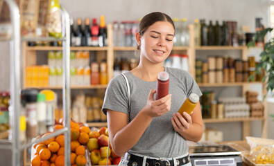 Youth girl spouses hold bottles and choose consider smoothie in store. Market visitor pick up cocktail made of fruits, berries, seeds. Client examines contents of package, chooses smoothie
