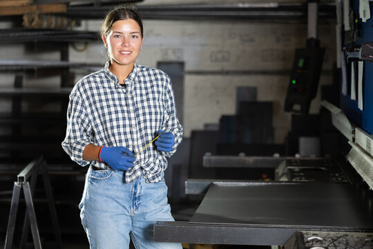 Young girl, professional technician with tape measure in hands, wearing checkered shirt and gloves, posing in industrial metal workshop, smiling confidently and friendly at camera..