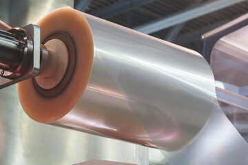 Roll of plastic film is being made in a factory. The roll is being fed into a machine that is cutting it into smaller pieces. The machine is surrounded by a metal frame