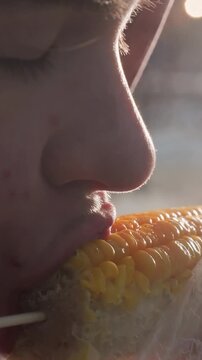close-up of person eating corn held by stick wrapped in white plastic with bokeh lights in blurred background, steam rising from fresh corn, enjoying tasty outdoor snack