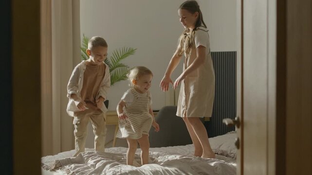 Three young children are joyfully jumping on a bed in a sunlit bedroom, full of laughter and playfulness.