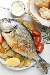 Tasty roasted dorado fish served with vegetables and sauce on white wooden table, flat lay. Homemade seafood dish