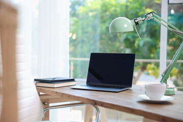Home workspace. Laptop, cup of drink, lamp and stationery on wooden desk near window indoors